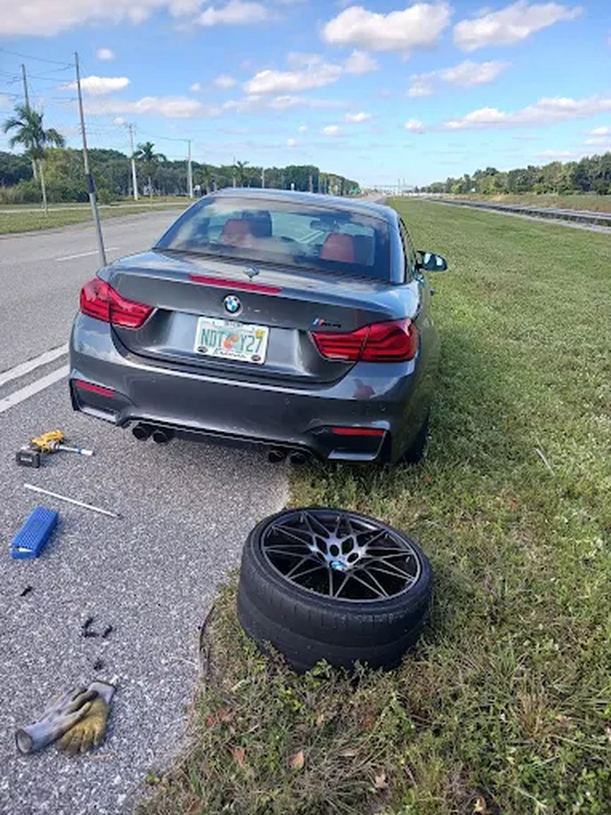 Grey BMW on the side of the road with a wheel off and the tire on the grass